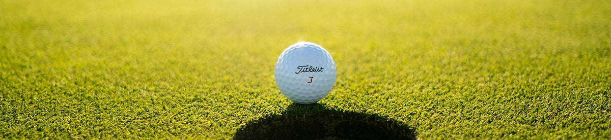 A white Titleist golf ball resting on a green grass surface near the hole. A typical Gripit Golf shot, with a warm golden light background.